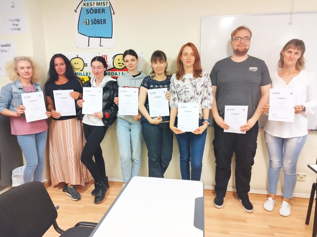 Группа студентов, мужчины и женщины, держат в руках дипломы об успешном окончании курса эстонского языка уровня А2 в классе учебного заведения Atlasnet в Эстонии, Таллинне.